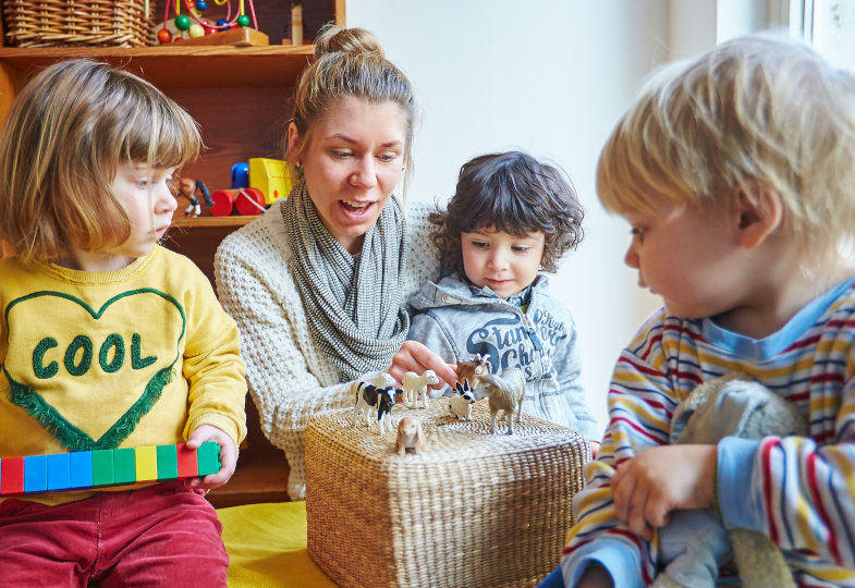 Erzieherin sitzt mit drei Kindern um einen umgestülpten Korb herum. Auf dem Korb stehen verschiedene Tierfiguren.