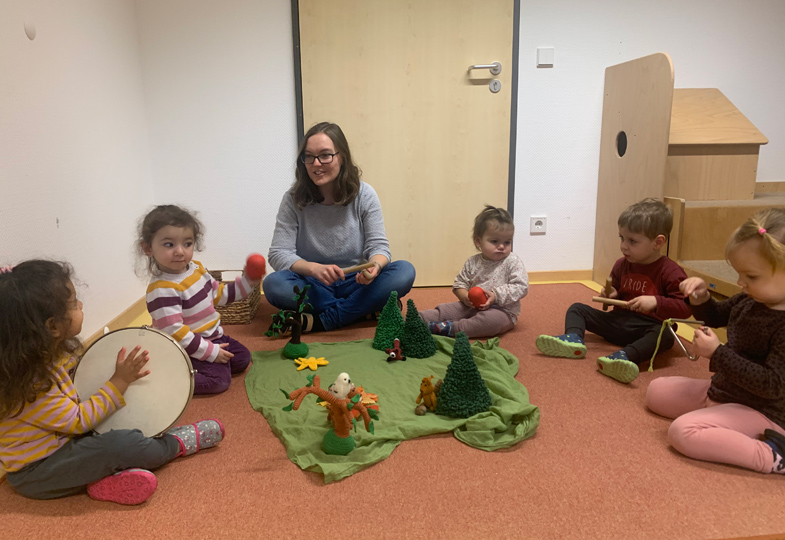 Kinder sitzen mit Musikinstrumenten im Kreis, in dessen Mitte ein "Wald" aufgebaut ist.